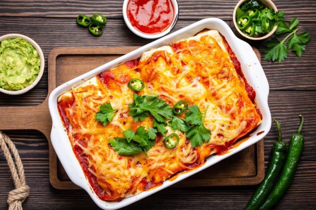 enchiladas with meat, chili red sauce and cheese in white casserole dish over rustic wooden background, served with guacamole and tomato dips